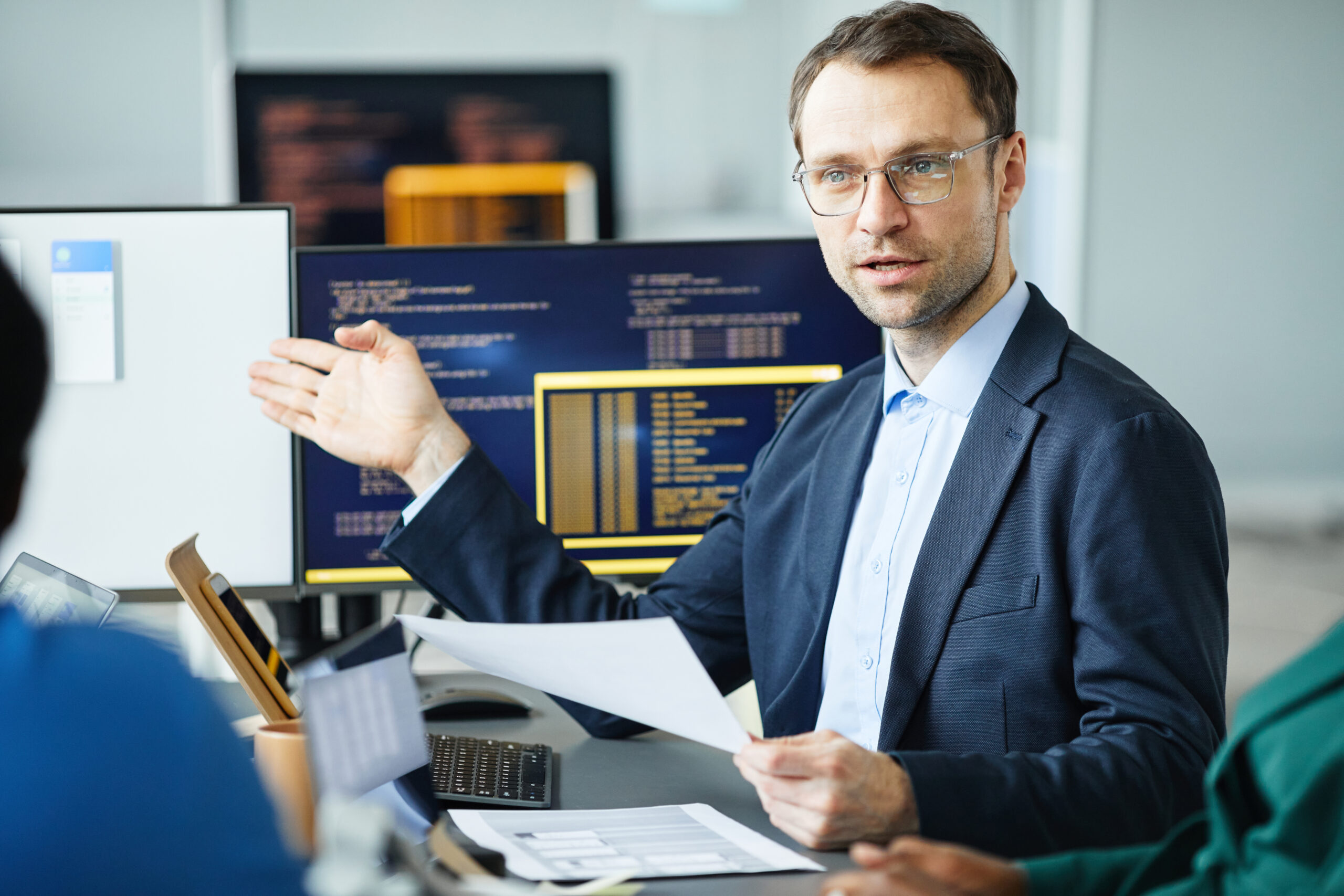 Mature developer pointing at computer monitor with interface and giving report to his colleagues at meeting